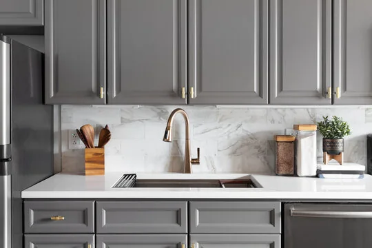 Gray raised-panel cabinets with brass pulls, a brass faucet and a marble-look backsplash